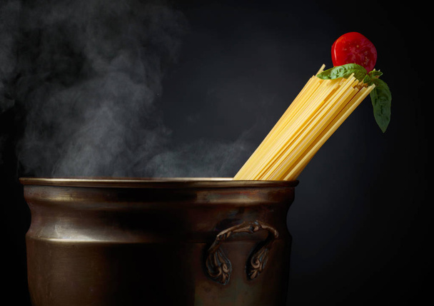 Raw spaghetti with tomato and basil in old brass pan with hot water. Black background. Copy space. - Photo, Image