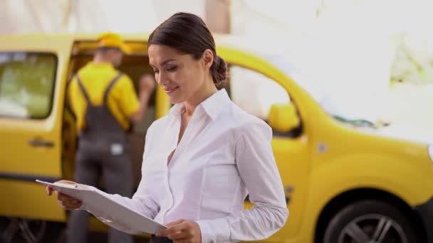 Smiling girl reads delivery papers on clipboard. Courier near the yellow car - Filmmaterial, Video