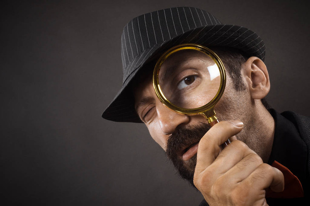 Staromódní detektiv s kloboukem se dívá přes Lupa nebo Lupa na tmavě šedém pozadí. - Fotografie, Obrázek