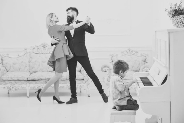 Boy adorable try to play piano musical instrument, while parents dancing. Child sit next to piano, play music, white interior background. Rich parents enjoy parenthood. Musician education concept - Фото, зображення
