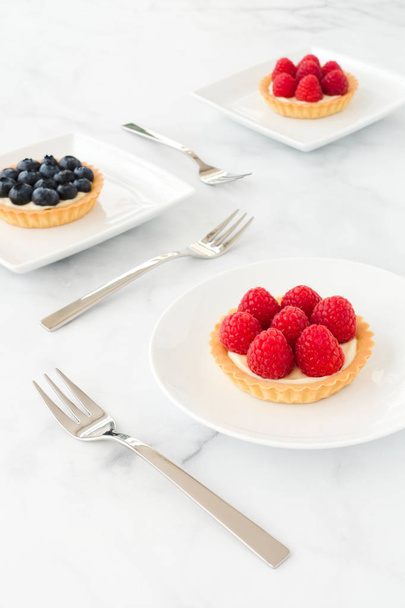 Raspberry and blueberry tartlets filled with vanilla cream on white porcelain plates and white marble. - 写真・画像
