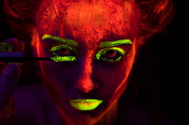 Обличчя красивої молодої сексуальної дівчини з ультрафіолетовою фарбою на її шкірі, яка малює вії пензлем у яскравій фарбі. Красива жінка з сяючим бодіарт в чорному ламповому світлі
 - Фото, зображення