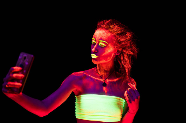 Una hermosa joven sexy con pintura ultravioleta en su cuerpo bailando en la oscuridad y haciendo selfie. Mujer bonita con el bodyart brillante en la luz de la lámpara negra
 - Foto, Imagen