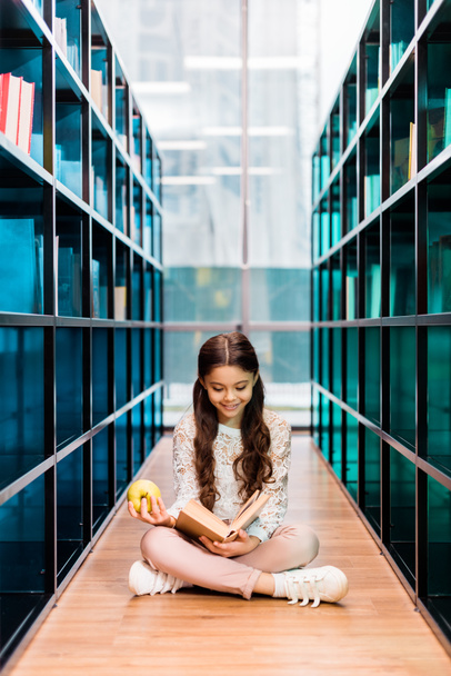 Urocza dziewczyna szczęśliwy trzyma jabłko i czytanie książki w bibliotece  - Zdjęcie, obraz