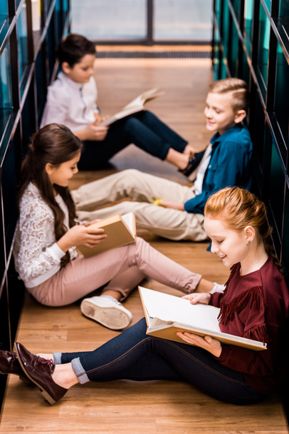 high angle view of four schoolchildren sitting on floor and reading books in library  - Фото, изображение