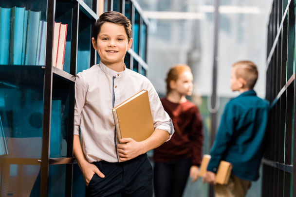 adorable boy holding book and smiling at camera while visiting library with classmates  - Photo, Image