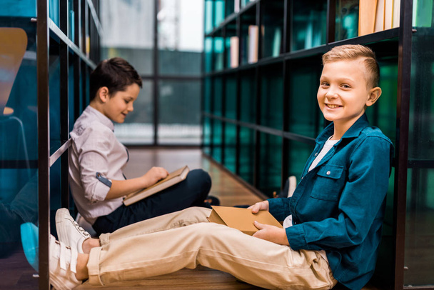 side view of cute smiling schoolboys reading books while sitting on floor in library   - Foto, afbeelding