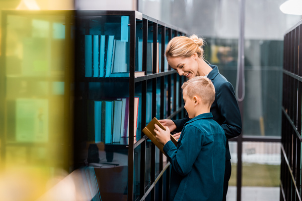 улыбающаяся молодая библиотекарша смотрит на школьника, держащего книгу в библиотеке
  - Фото, изображение