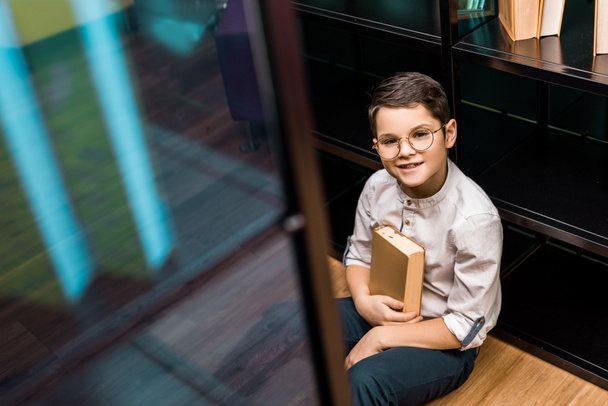high angle view of smiling schoolboy in eyeglasses holding book and sitting on floor in library   - Foto, Imagen