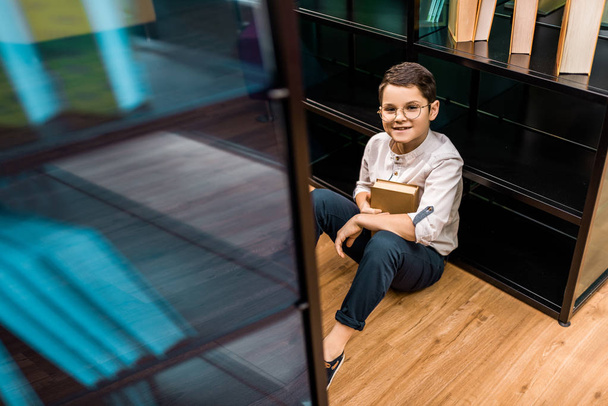 high angle view of schoolboy in eyeglasses holding book and smiling at camera in library   - Foto, afbeelding