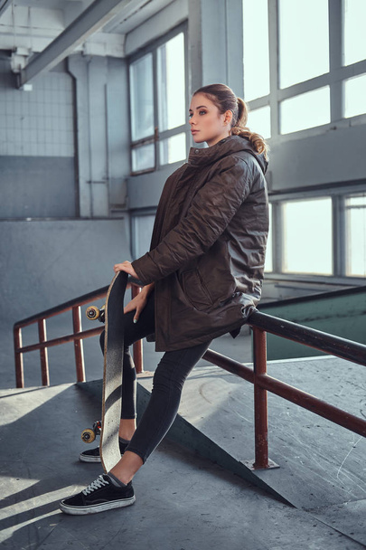 Een mooi jong meisje draagt een geruite hemd en jas met haar skateboard zittend op een spoor van de sleur in skatepark binnenshuis. - Foto, afbeelding