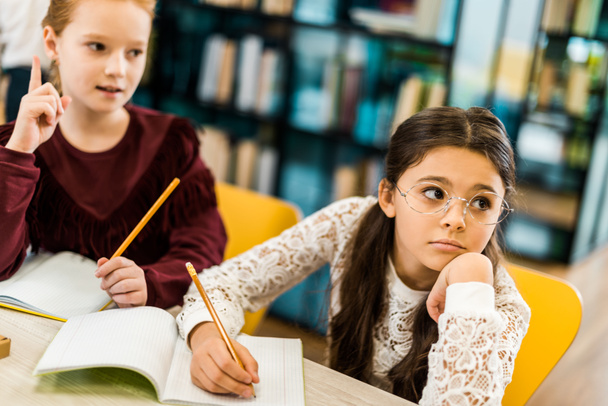 entzückende Schulmädchen, die mit Bleistiften schreiben und wegschauen, während sie in der Bibliothek lernen  - Foto, Bild