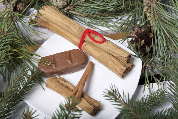 Cinnamon sticks and chocolate next to the pine twigs and cones. On a wooden box. - Фото, изображение