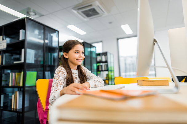 чарівна усміхнена школярка використовує настільний комп'ютер у бібліотеці
   - Фото, зображення