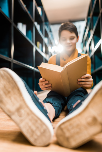 selective focus of schoolboy reading book and sitting on floor in library     - Фото, изображение