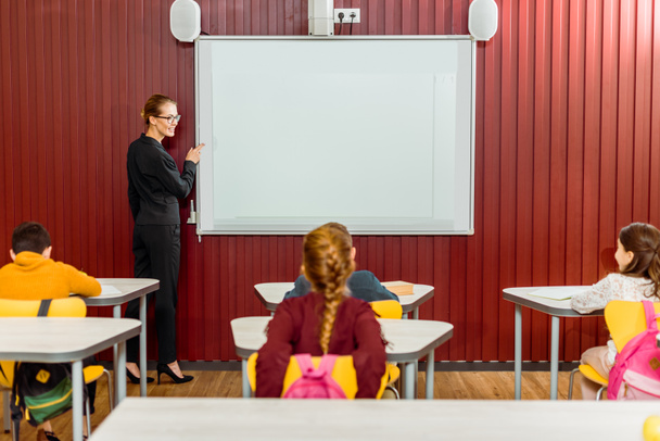 smiling teacher pointing at interactive whiteboard and looking at schoolkids - Photo, Image