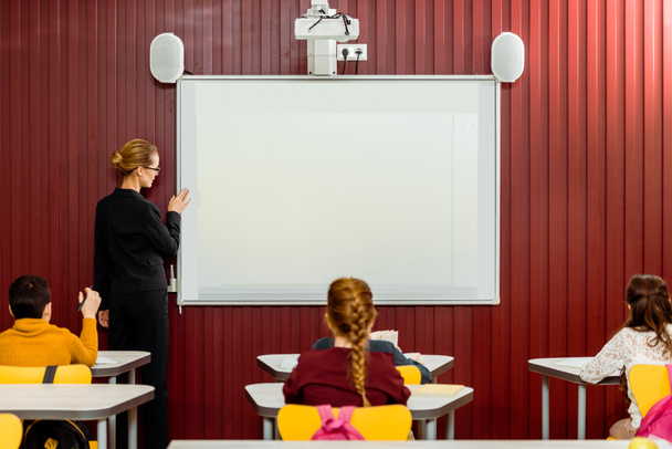 vue arrière des écoliers assis aux bureaux et de l'enseignant faisant la présentation au tableau blanc
  - Photo, image