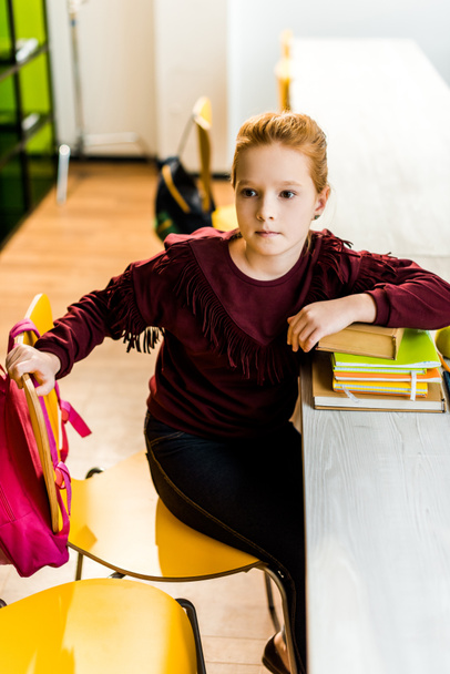 adorable schoolkid sitting at desk with books and looking away in library - Foto, Bild