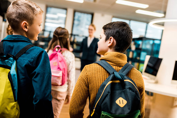 visão traseira de meninos com mochilas olhando uns para os outros ao visitar biblioteca com colegas de classe
 - Foto, Imagem
