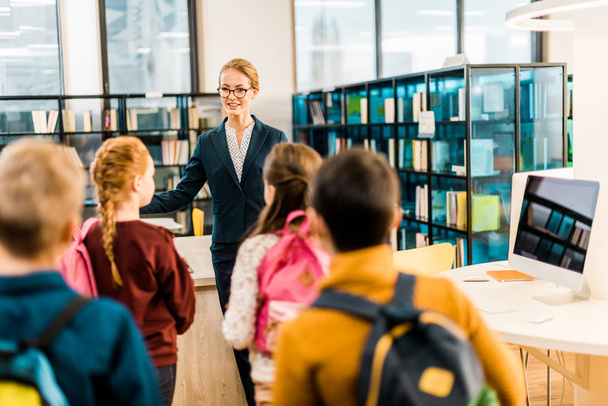 Achteraanzicht van schoolkinderen met rugzakken vrouwelijke bibliothecaris kijken  - Foto, afbeelding