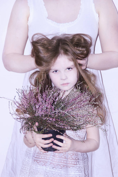 Μαμά και κόρη σε λευκό φορέματα σε άσπρο φόντο απεικονίζουν χειμώνα και την άνοιξη, κρατώντας λουλούδια και ένα κλαδί με φύλλα. Μαμά τα μαλλιά της κόρης κρατά στα χέρια της - Φωτογραφία, εικόνα