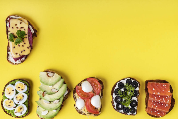 On the yellow background there are a few sendwiches with tomatoes, eggs, chees, avocado, fish, olives, ham. Top view. Horizontal orientation. - Photo, Image