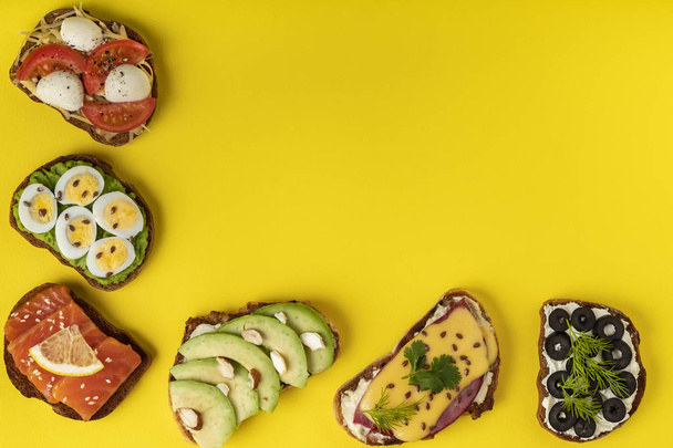 On the yellow background there are a few sendwiches with tomatoes, eggs, chees, avocado, fish, olives, ham. Top view. Horizontal orientation. - Photo, Image