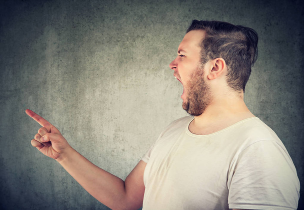 Νεαρός άνδρας casual παχουλός φωνάζει με θυμό και δείχνει δάχτυλο σε κάποιον σε γκρίζο φόντο - Φωτογραφία, εικόνα