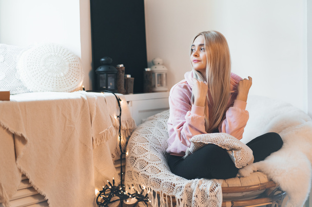 Cute teenage girl sitting in a comfy round armchair at home legs crossed. She wears a pale pink parka. Candles on the window in the background. - Фото, изображение