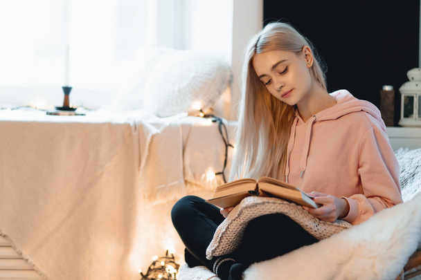 Young woman chilling at home in a comfortable round armchair near the window. Girl relaxing, reading and dreaming book in the loft living room. Imagining the book extraordinary world. - Fotó, kép
