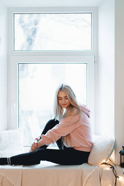 Lovely girl sitting on the windowsill with pillows around thinking and dreaming indoors. She has long blonde hair and a shy smile. - Фото, изображение