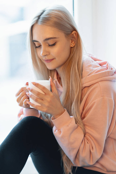 A pretty young girl enjoying a cup of coffee or milk while sitting by the window. Wearing a casual pink, pale parka. A cute young woman with elf alike facial features holding a white mug - Photo, Image