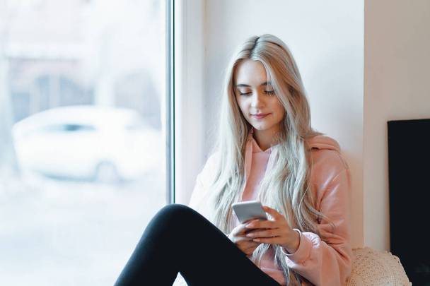 Pretty girl sitting on the window sill with smartphone in hands. She has long blonde hair, smile and looking at her phone. Wearing pink, pale parka. - Foto, imagen