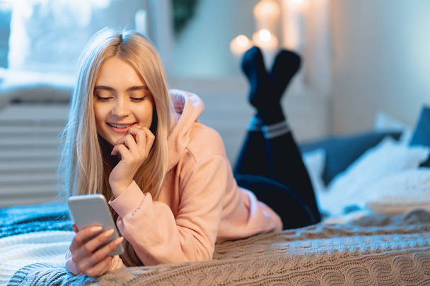 Smiling pretty blond girl in pink parka and black leggings using a smart phone lying on the bed at home, while chatting with her friends - Фото, зображення