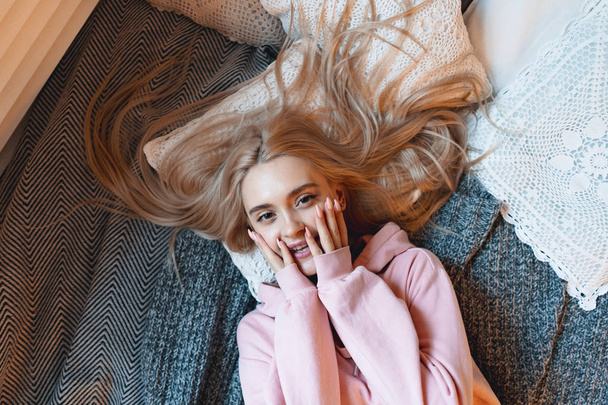 Beautiful smiling girl, with long blond hair, enjoying happiness, she cant belive its true. Portrait gorgeous young woman lying on the bad with pillows in comfy pink pale parka looking at camera. - Fotografie, Obrázek