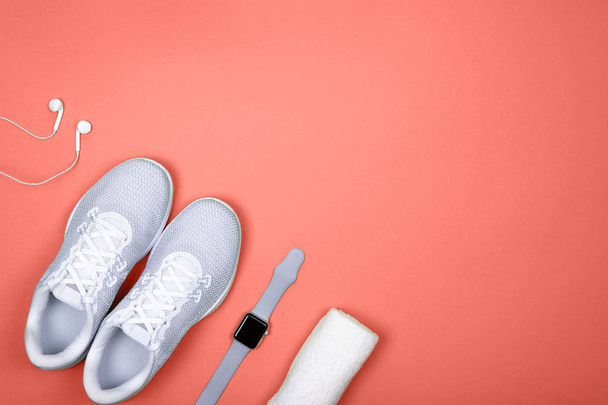 Λευκά αθλητικά παπούτσια (σπορτέξ) με έξυπνο ρολόι (δραστηριότητα tracker), ακουστικά (ακουστικά) και πετσέτα στο σαλόνι κοραλί χρώμα φόντου.   - Φωτογραφία, εικόνα