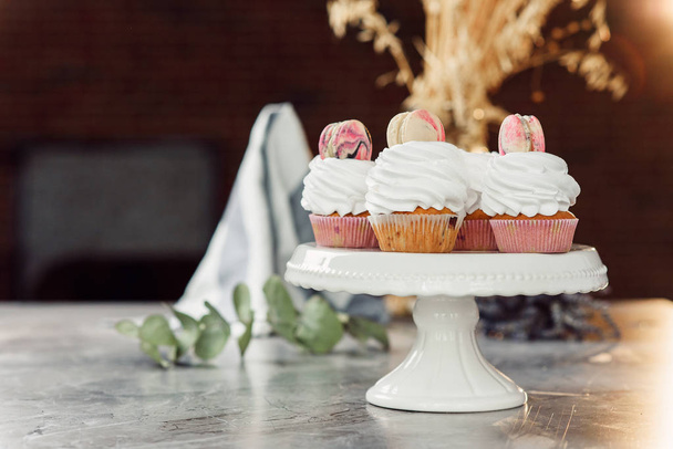 Fresh delicious cupcakes with white cream and colored macarons on a white tray on marble table. - Photo, Image
