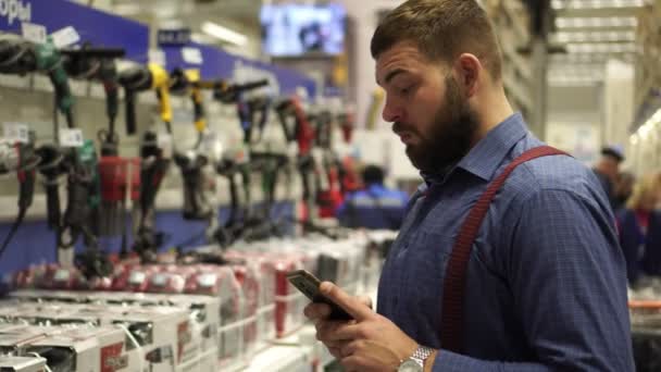 ενήλικα άνδρα σε ένα κατάστημα υλικού επιλέγει εργαλεία και χρησιμοποιεί ένα smartphone. - Πλάνα, βίντεο