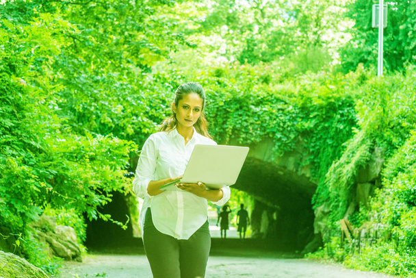 junge ostindische Amerikanerin auf Reisen, arbeitet in New York, hält Laptop in der Hand, geht im Central Park spazieren, guckt. Straßenbrücke mit grünen Blättern im Hintergrund. Farbe Ufo grün gefiltertes Klo - Foto, Bild