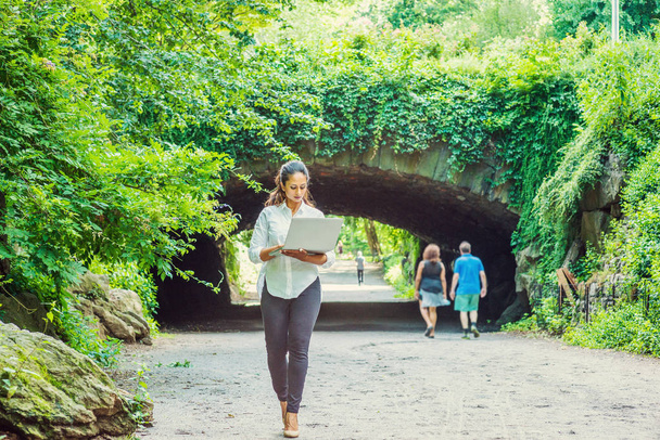 Young East Indian American Woman traveling, working in New York, wearing white shirt, black pants, holding laptop computer, walking at Central Park. Street bridge with green leaves on background - Photo, Image
