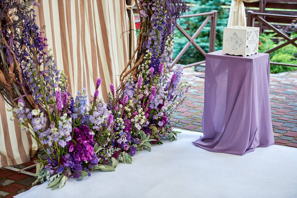 Prachtige cirkel huwelijksboog versierd met violette bloemen en bladeren buiten en tabel met paarse doek, kopie ruimte. Decoraties voor de ceremonie van het huwelijk in de open lucht - Foto, afbeelding