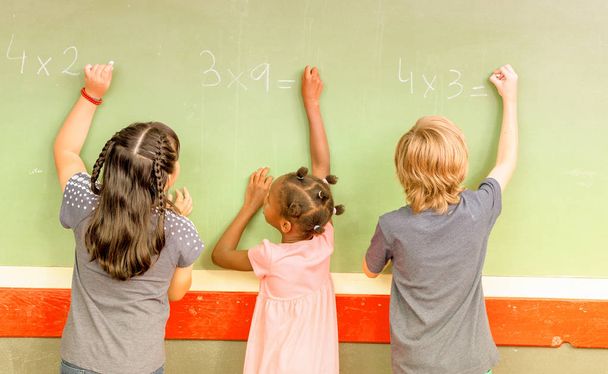 Dzieci w wieku szkolnym mieszanej rasy pisania na tablicy. - Zdjęcie, obraz