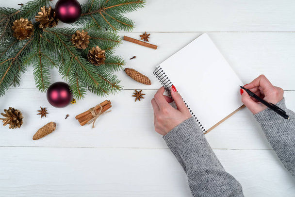 Top view of Christmas background with blank empty diary and girl's hands with pen writing goals on white wooden background, free space. Flat lay, top view - Zdjęcie, obraz