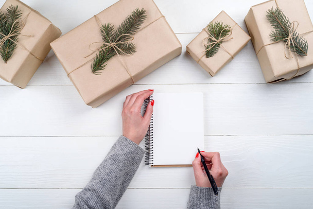 Christmas background with blank empty notebook and girl's hands with pen writing goals on white wooden background, free space. Flat lay, top view. Holiday gift boxes wrapped in kraft - Φωτογραφία, εικόνα