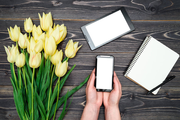Bouquet of yellow tulips, tablet, open empty notebook and cellphone with white blank screen in woman hands on wooden background, copy space. Top view, flat lay. Womens Day, Mothers day, Easter  - Fotografie, Obrázek