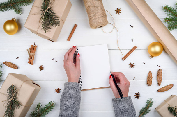 Top view of Christmas background with blank empty diary and girl's hands with pen writing goals on white wooden background, free space. Flat lay. Holiday gift boxes wrapped in kraft - Φωτογραφία, εικόνα