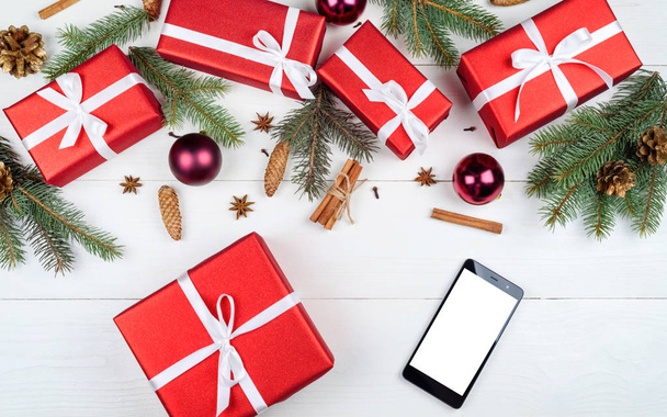 Christmas background with red gift boxes, fir tree branches, cones, baubles and mobile smart phone with white blank screen for app presentation, copy space. Top view, flat lay  - Φωτογραφία, εικόνα