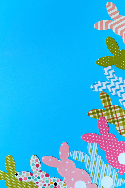 Easter bunny decoration on blue background, copy space. DIY holiday handicraft of colorful rabbits. Flat lay, top view. Border of paper rabbits cutouts. Easter greeting card. Happy Easter concept - 写真・画像