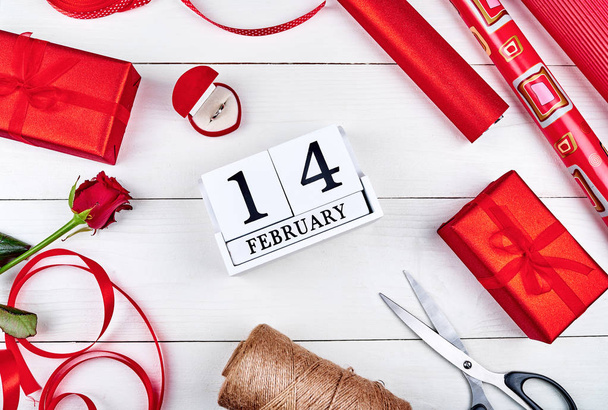 Gift wrapping background. Rolls of red wrapping paper, ribbon, red rose, giamond ring, gift boxes and wood calendar february 14 on wooden background, free space. Flat lay, top view  - Foto, immagini