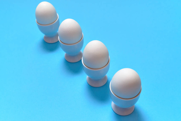 Valkoiset munat seisoo muna kuppi eristetty sinisellä pohjalla, kopioi tilaa. Keitettyjen munien rivi jalustalla. Terveellistä ruokaa. Pääsiäismunia. Hyvää pääsiäistä käsite
 - Valokuva, kuva